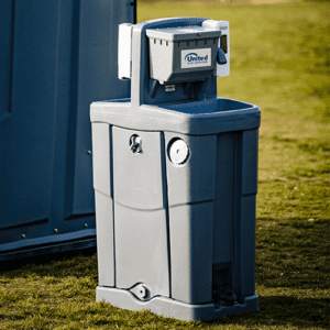 A portable gray handwashing station with a paper towel dispenser and a soap dispenser, set on a grassy area beside a blue portable toilet